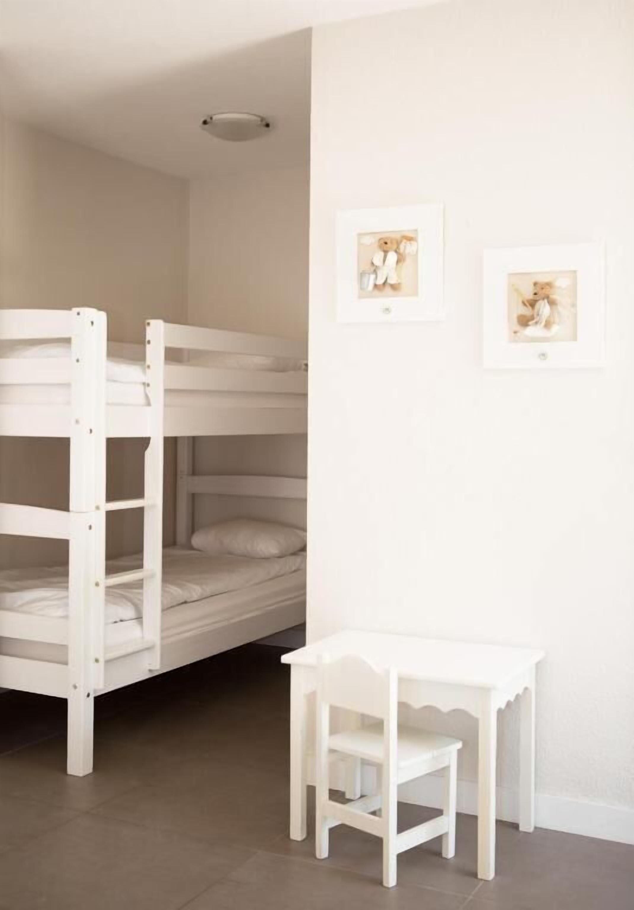 Una cama litera con estructura blanca y una pequeña mesa blanca con una silla en una habitación con paredes lisas y una luz en el techo.