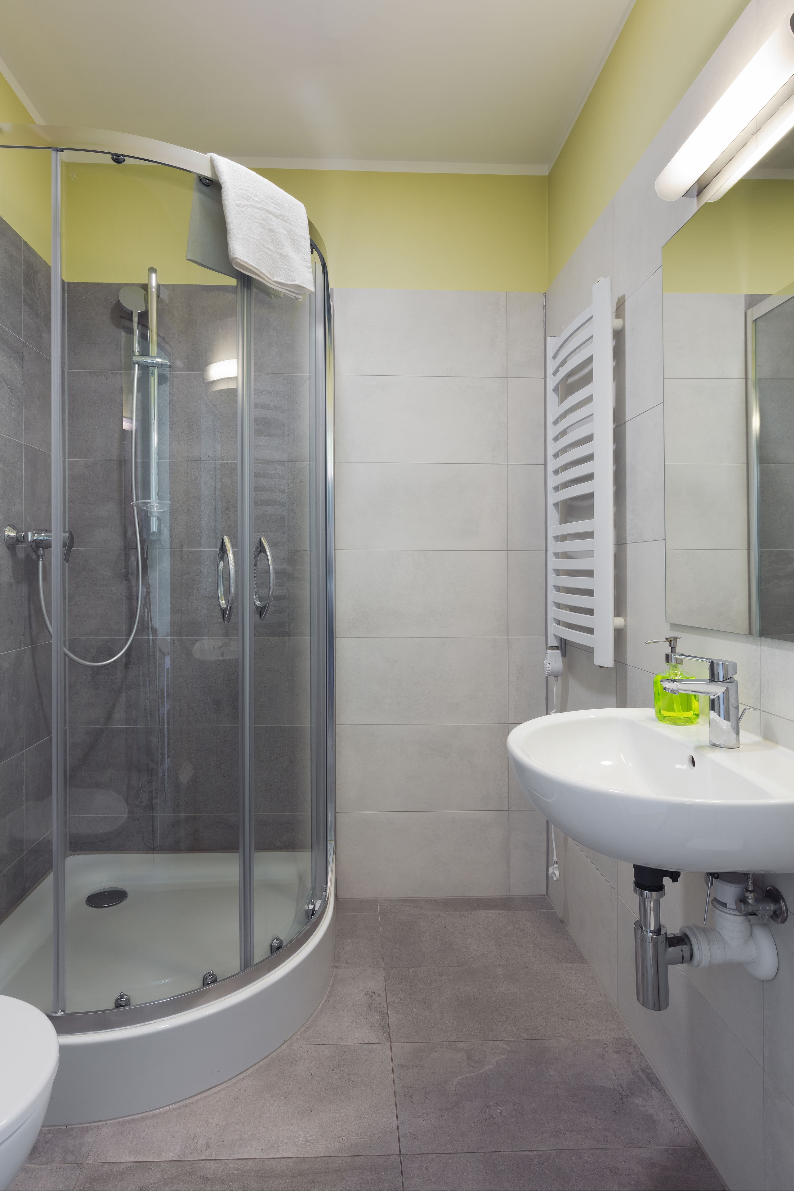 A modern bathroom with a glass-enclosed shower, a white sink with a green soap dispenser, and a white towel hanging on the shower door.