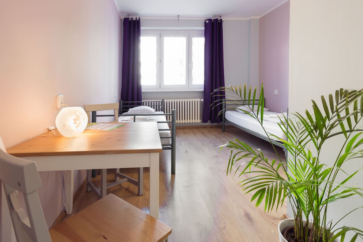 A small, well-lit room with a wooden table, a chair, a bed, and a potted plant.