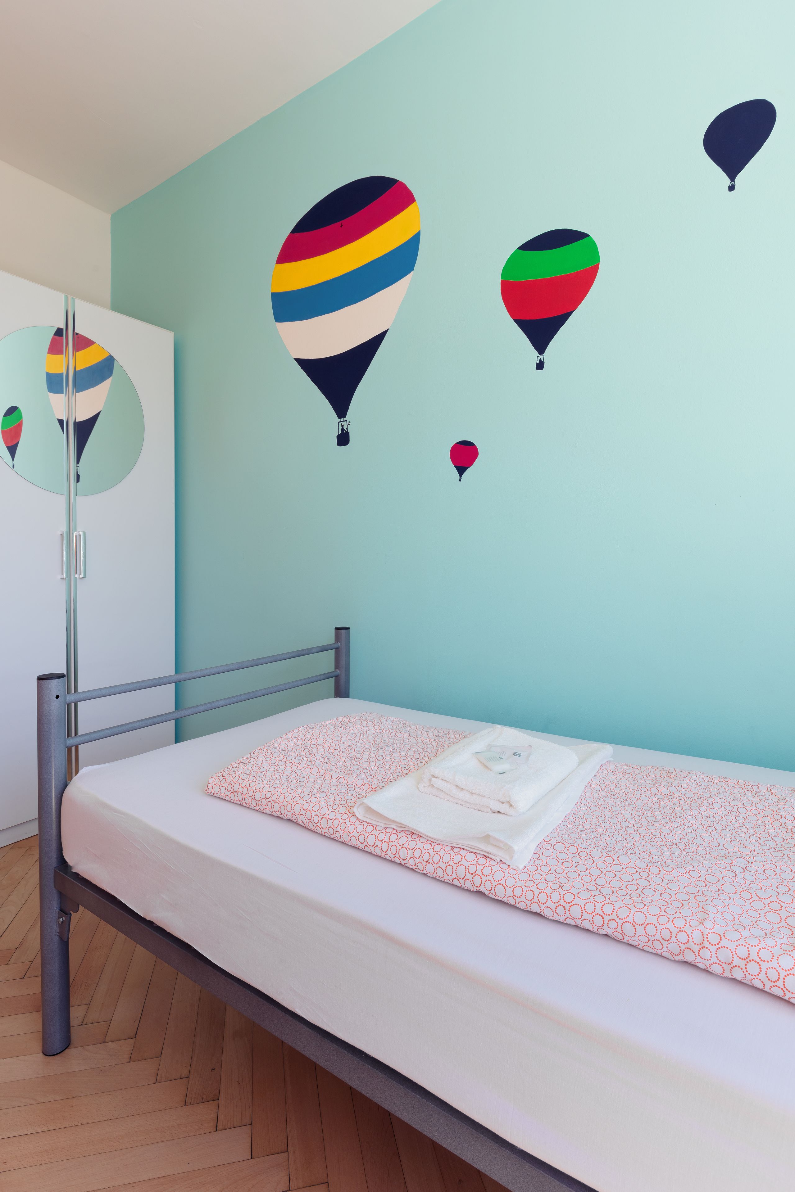 Ein Schlafzimmer mit einem Metallbettgestell, einer weißen Matratze und einer hellblauen Wand. An der Wand befinden sich drei Heißluftballons.