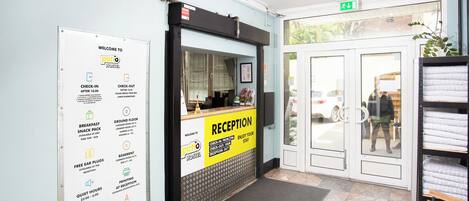 Un área de recepción con mostrador de check-in, escritorio de recepción y un cartel que detalla comodidades como aperitivos para el desayuno, tapones para los oídos gratuitos, horario de silencio y acceso a Wi-Fi.