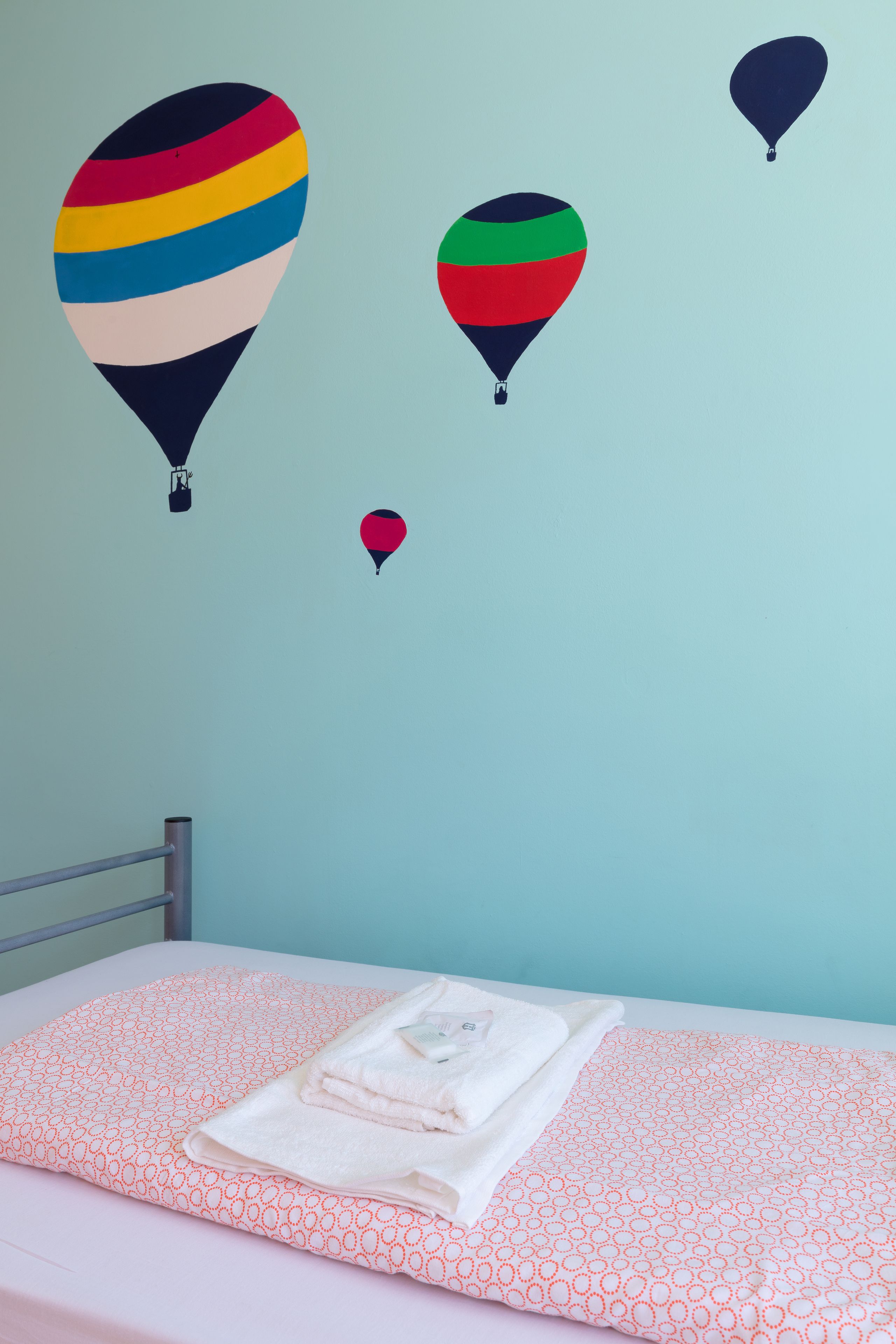 Een bed met een roze en oranje beddegoed met patroon, een keurig opgevouwen witte handdoek, en een muur met drie luchtballonstickers.