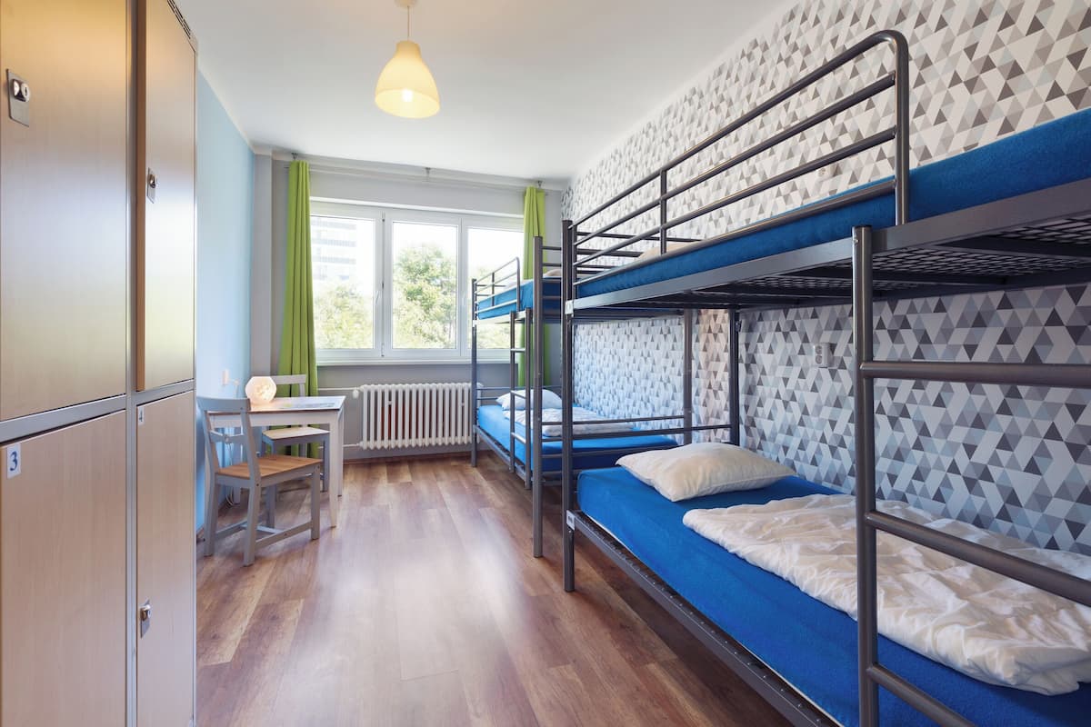 A dormitory room with bunk beds, a desk, and a window with curtains.