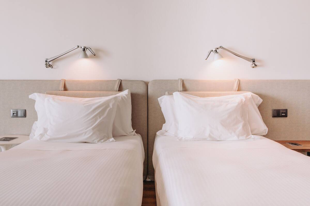 Een hotelkamer met twee bedden, elk met een nachtkastje en lamp, en een wandbeugelthermostaat.