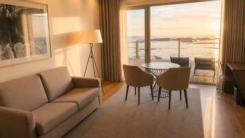 Une chambre d’hôtel avec un canapé, une table avec des chaises, une lampe sur pied et une grande fenêtre donnant sur la mer.