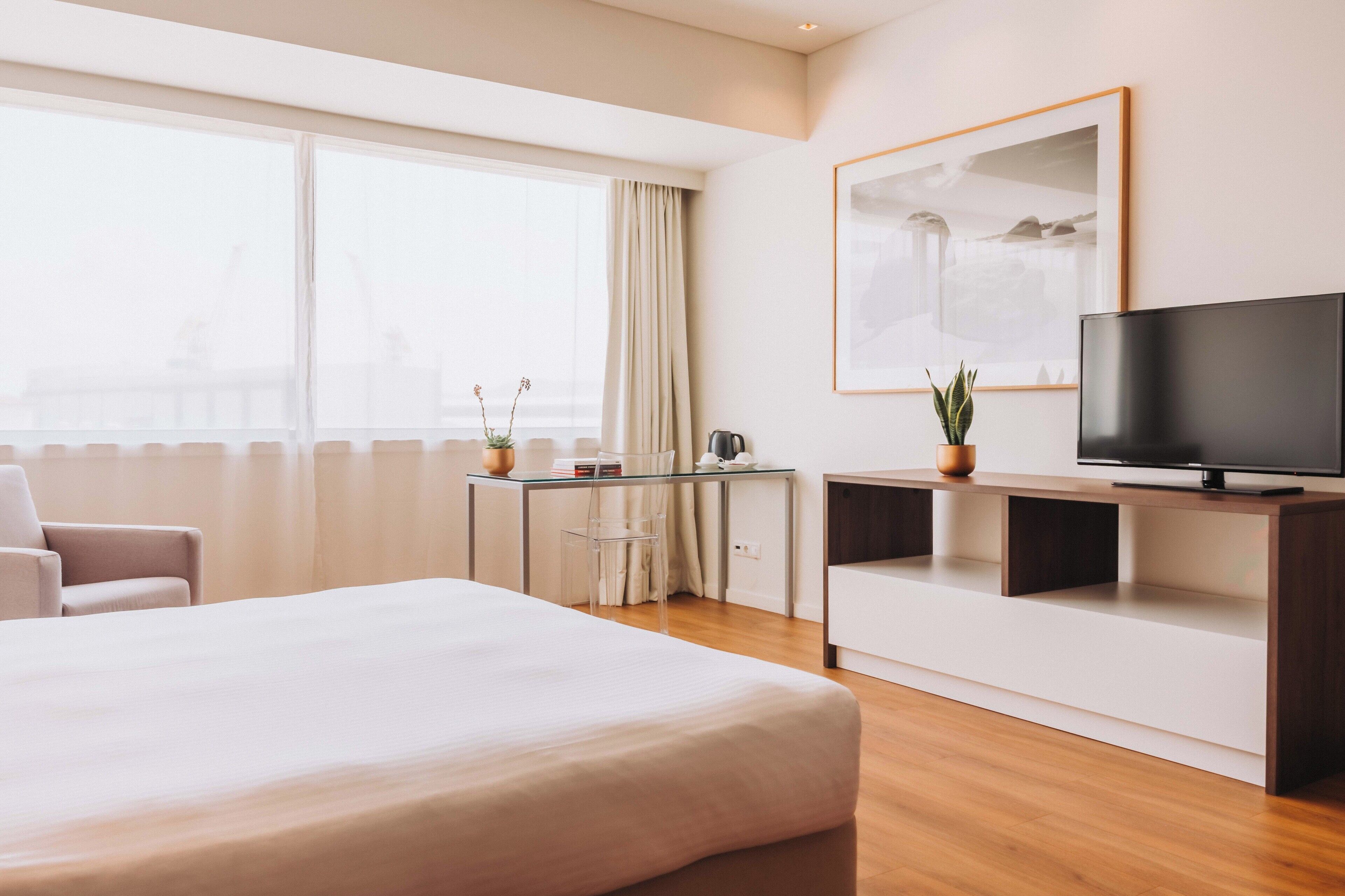 Habitación de hotel con una cama grande, un escritorio con silla, televisión y ventana con cortinas.