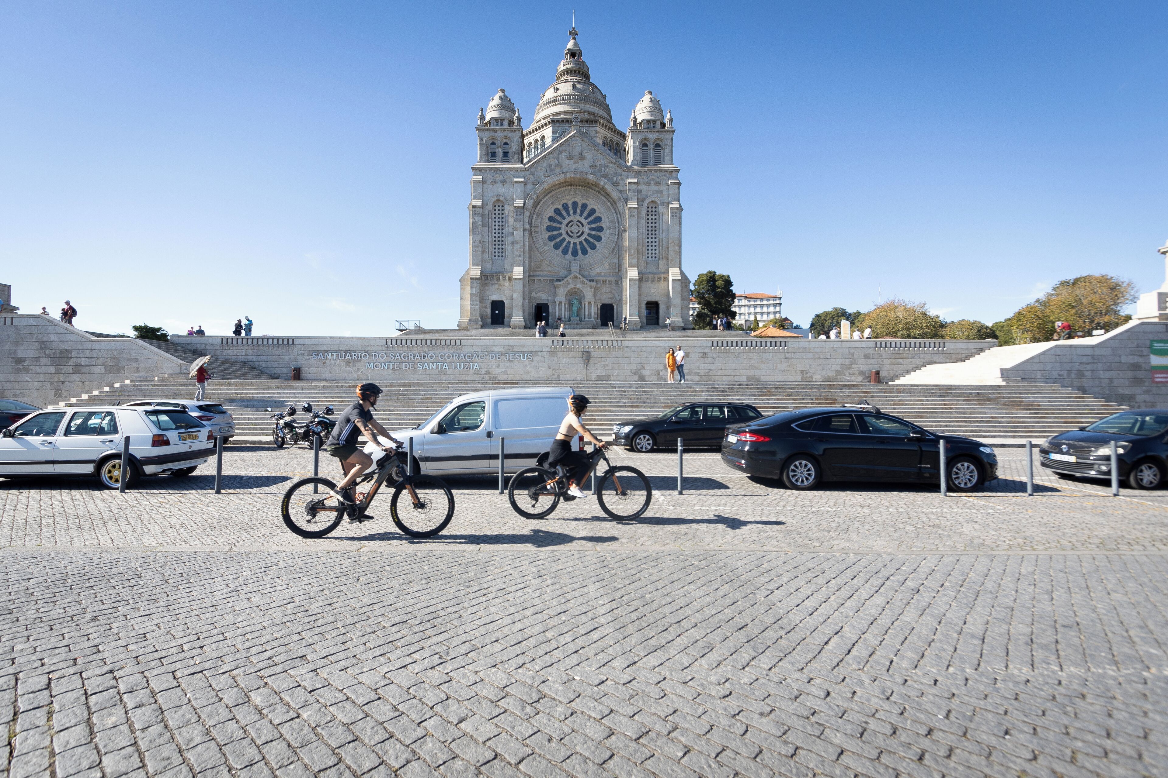 A large cathedral with a prominent circular window and two towers, surrounded by a paved area with cars and bicycles.