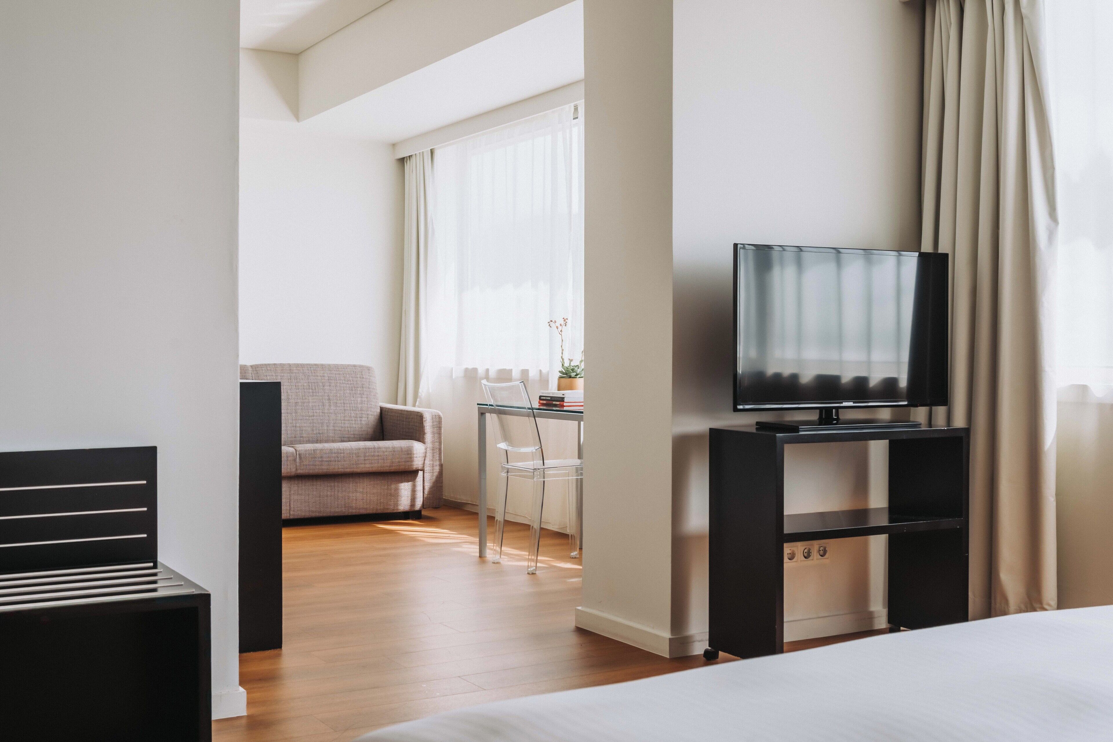 Une chambre d’hôtel avec un lit, un téléviseur posé sur un meuble, une chaise, une petite table avec une plante et une fenêtre avec des rideaux.