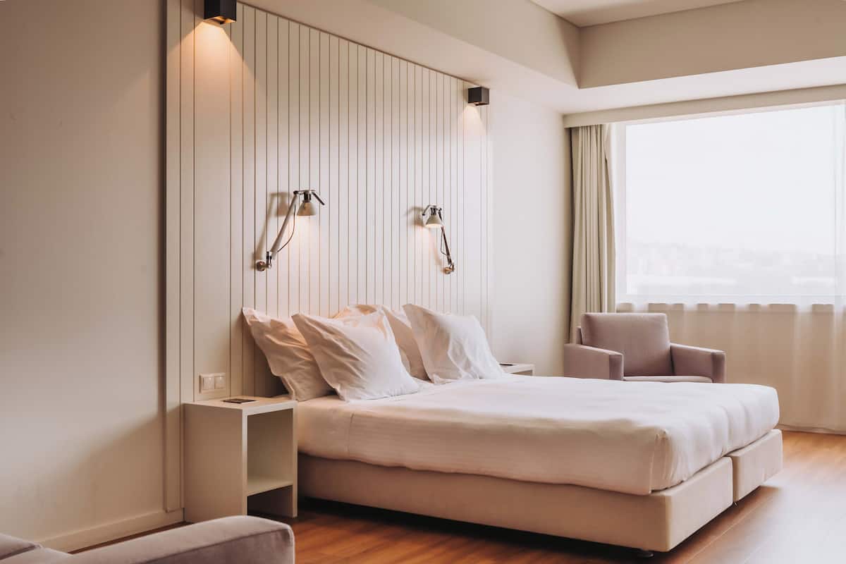Een moderne slaapkamer met een groot bed, nachtkastjes, twee wandlampen, een raam met gordijnen en uitzicht op de stad.