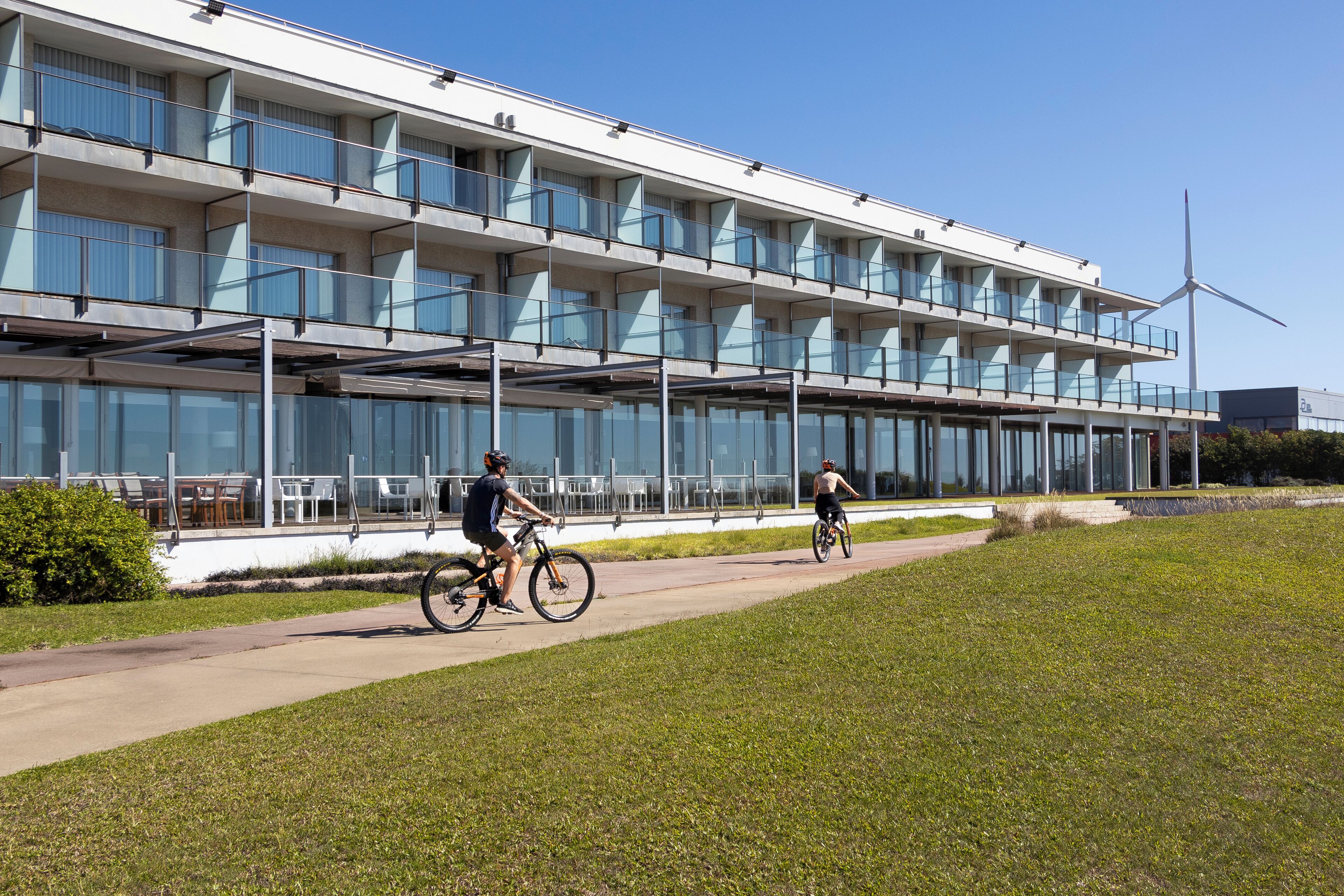 A modern building with multiple balconies and large windows, a pathway, and a wind turbine in the background.