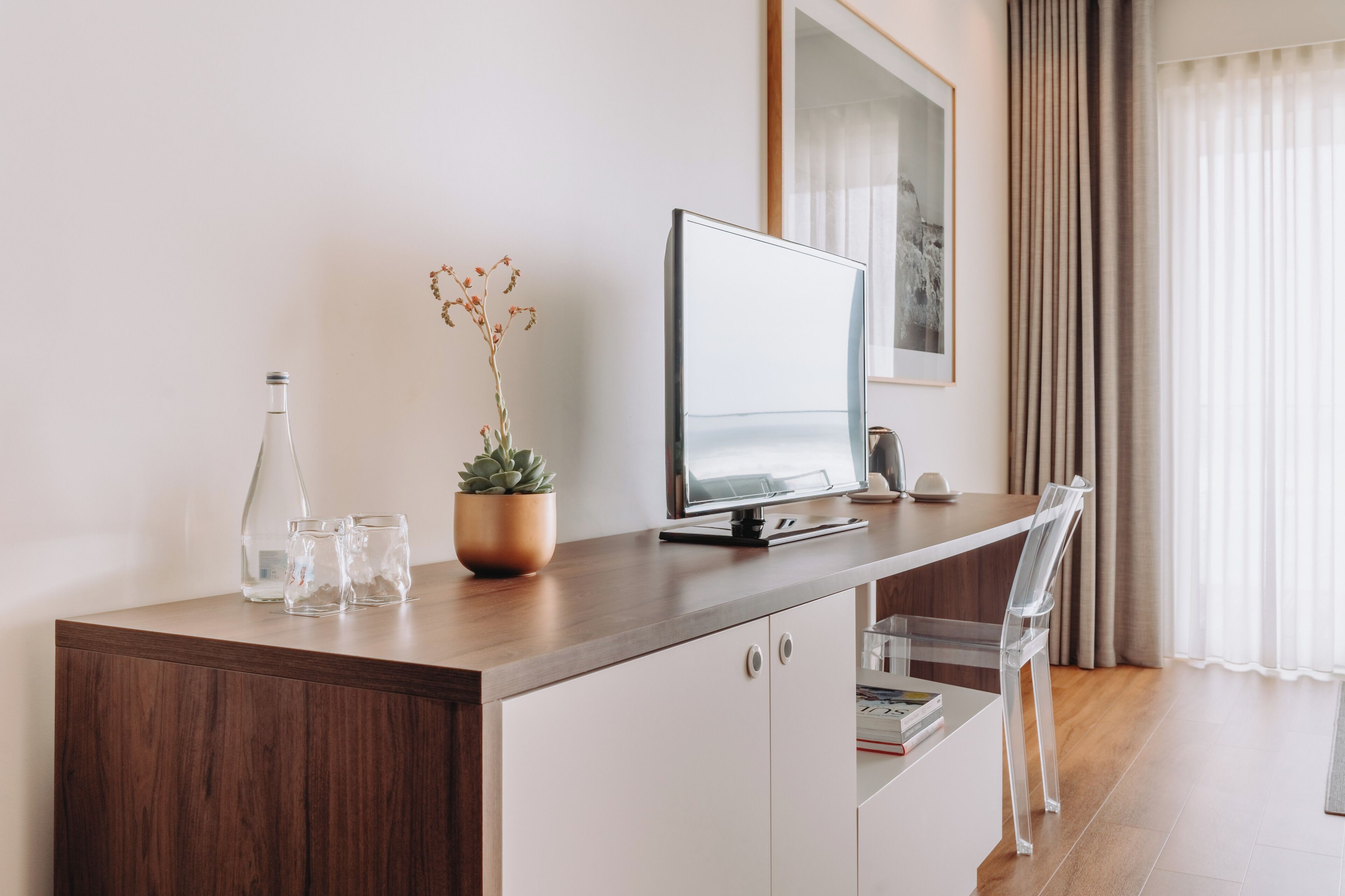 A modern hotel room with a wooden desk, a computer, a potted plant, a glass vase, and a clear chair.
