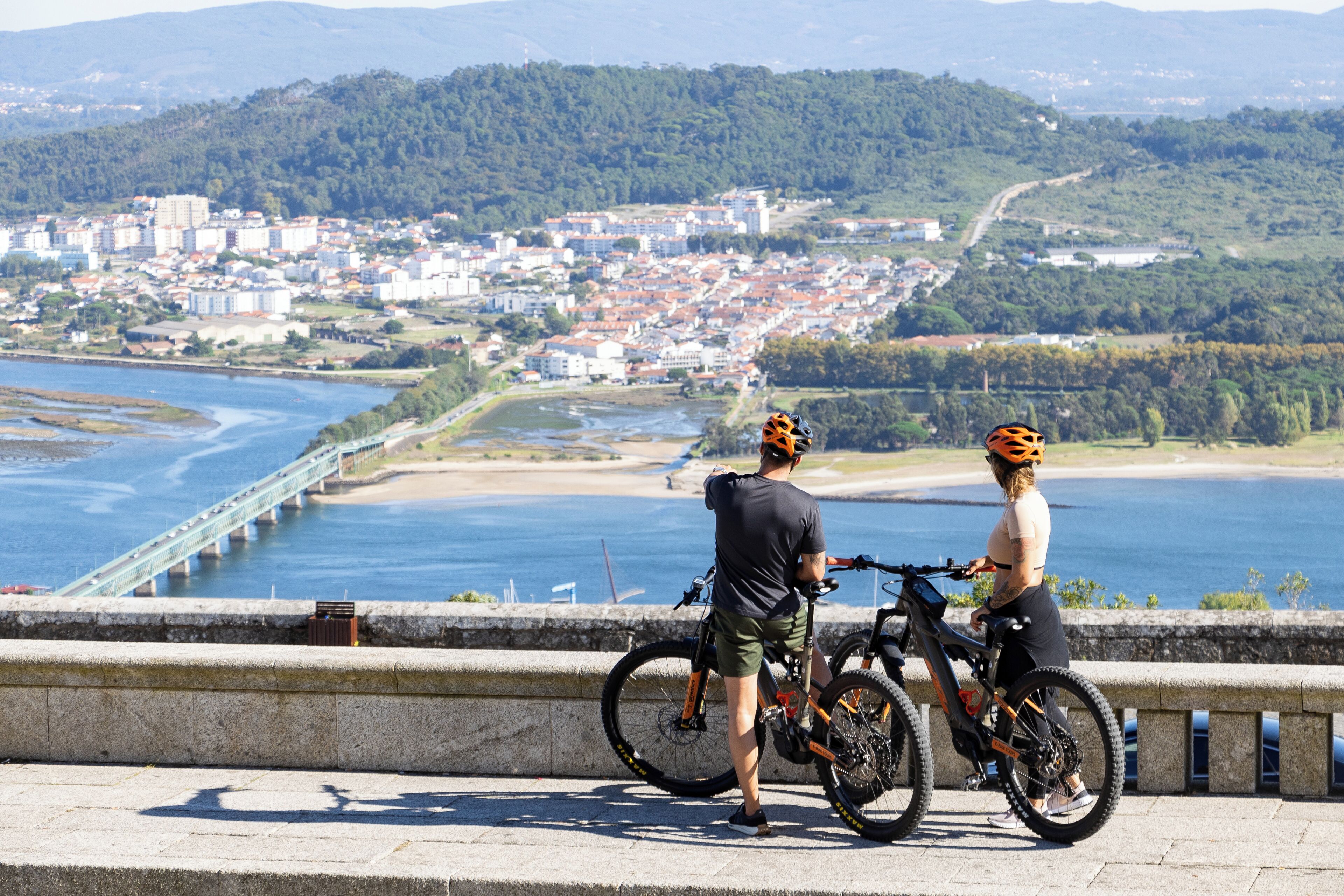 A coastal cityscape with a bridge, two cyclists, and a view of the sea and hills.