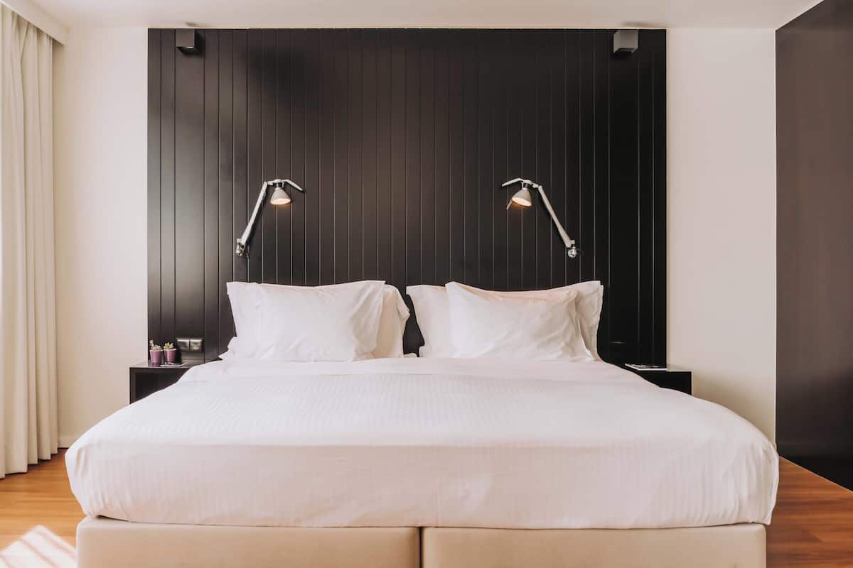 Een groot bed met twee bedlampen, een hoofdbord van donker hout en een klein nachtkastje met een plant.