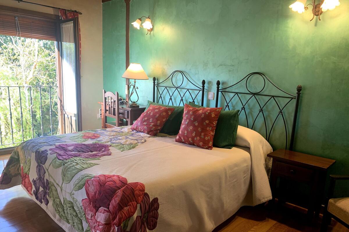 A bedroom with a bed, green walls, a wooden floor, a nightstand, a lamp, and a view of trees through a window.