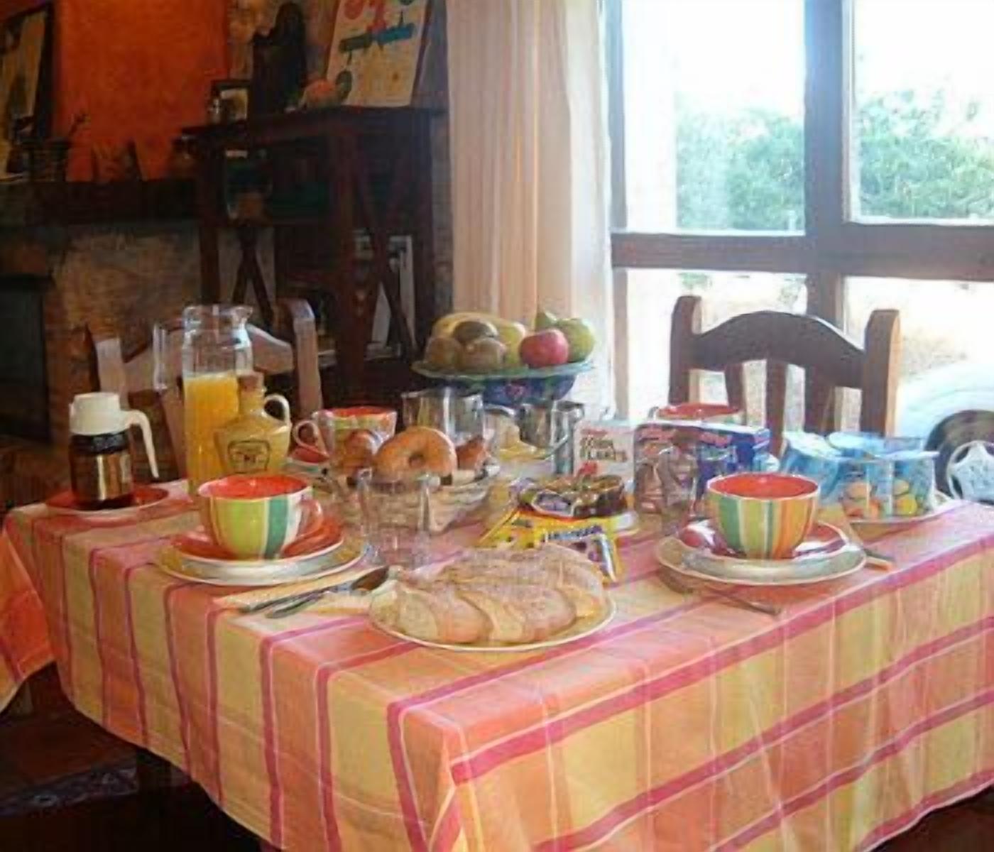 Une table de petit-déjeuner avec divers aliments et boissons, un pichet de jus d’orange et un saladier de fruits.