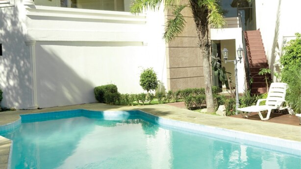 Una piscina rectangular con agua azul cristalina, rodeada por una terraza de piedra. Se observan sillas reclinables blancas y una pequeña palmera al fondo.