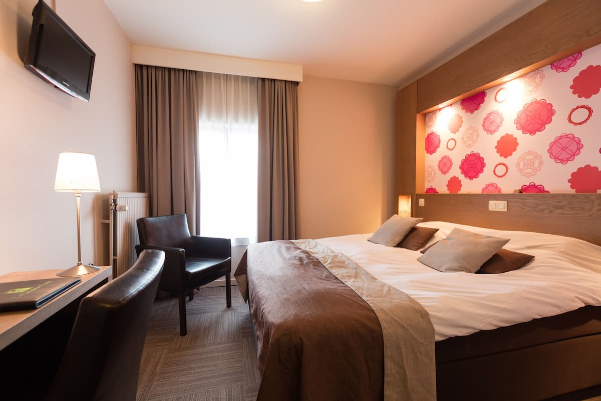 A hotel room with a bed, a desk with a laptop, a chair, a TV mounted on the wall, and a window with curtains.