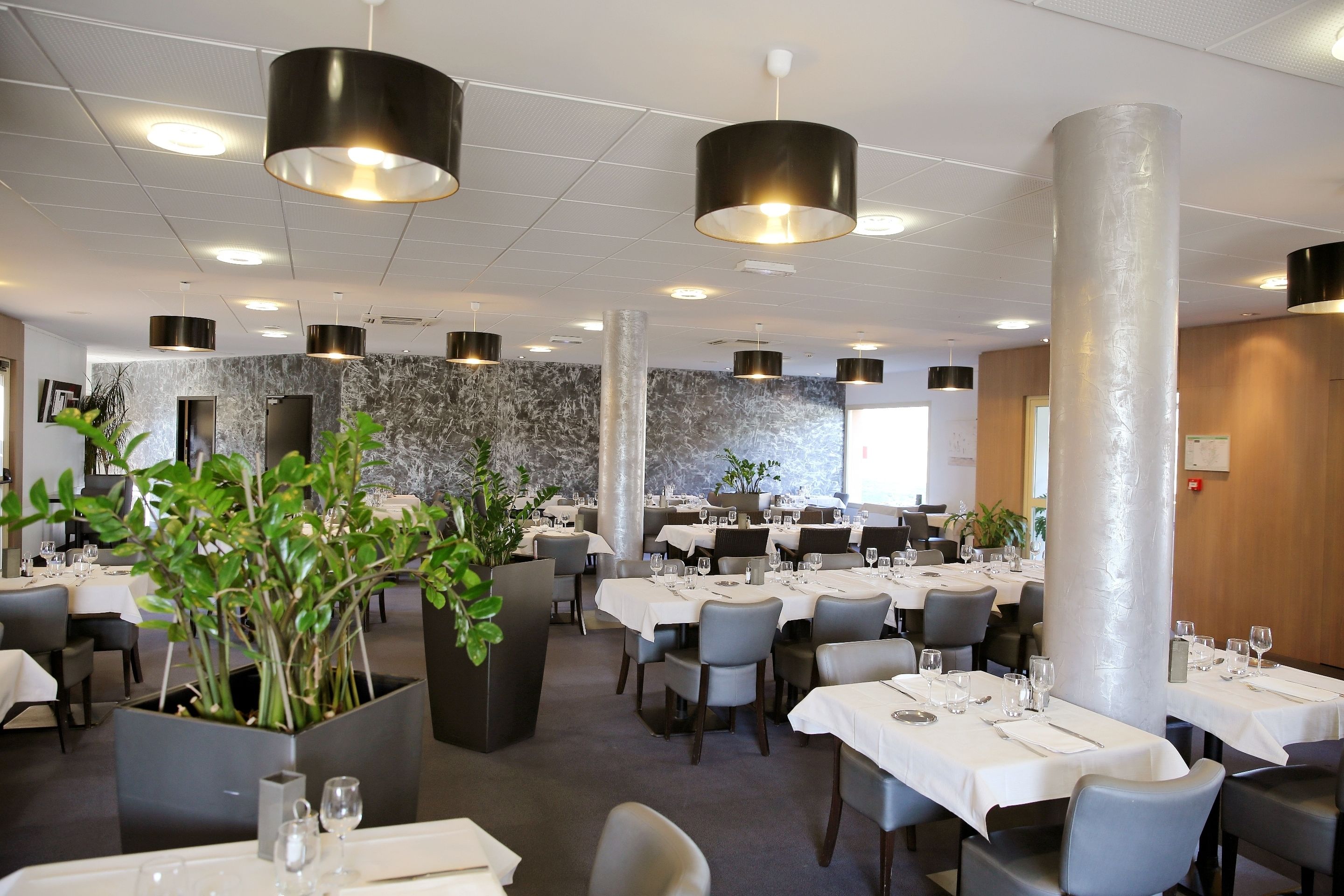 A restaurant with multiple tables, black pendant lights, marble walls, and potted plants.