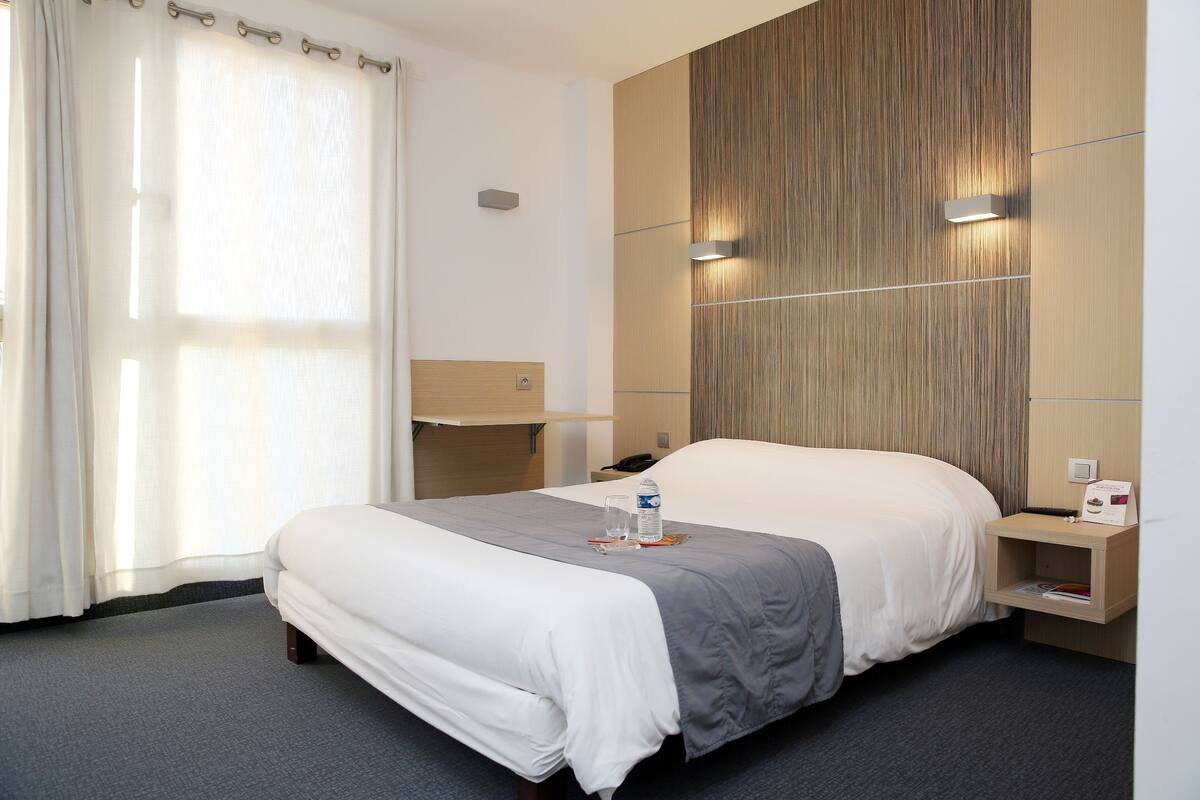Habitación de hotel con cama, mesitas de noche, lámpara de pared y ventana con cortinas.