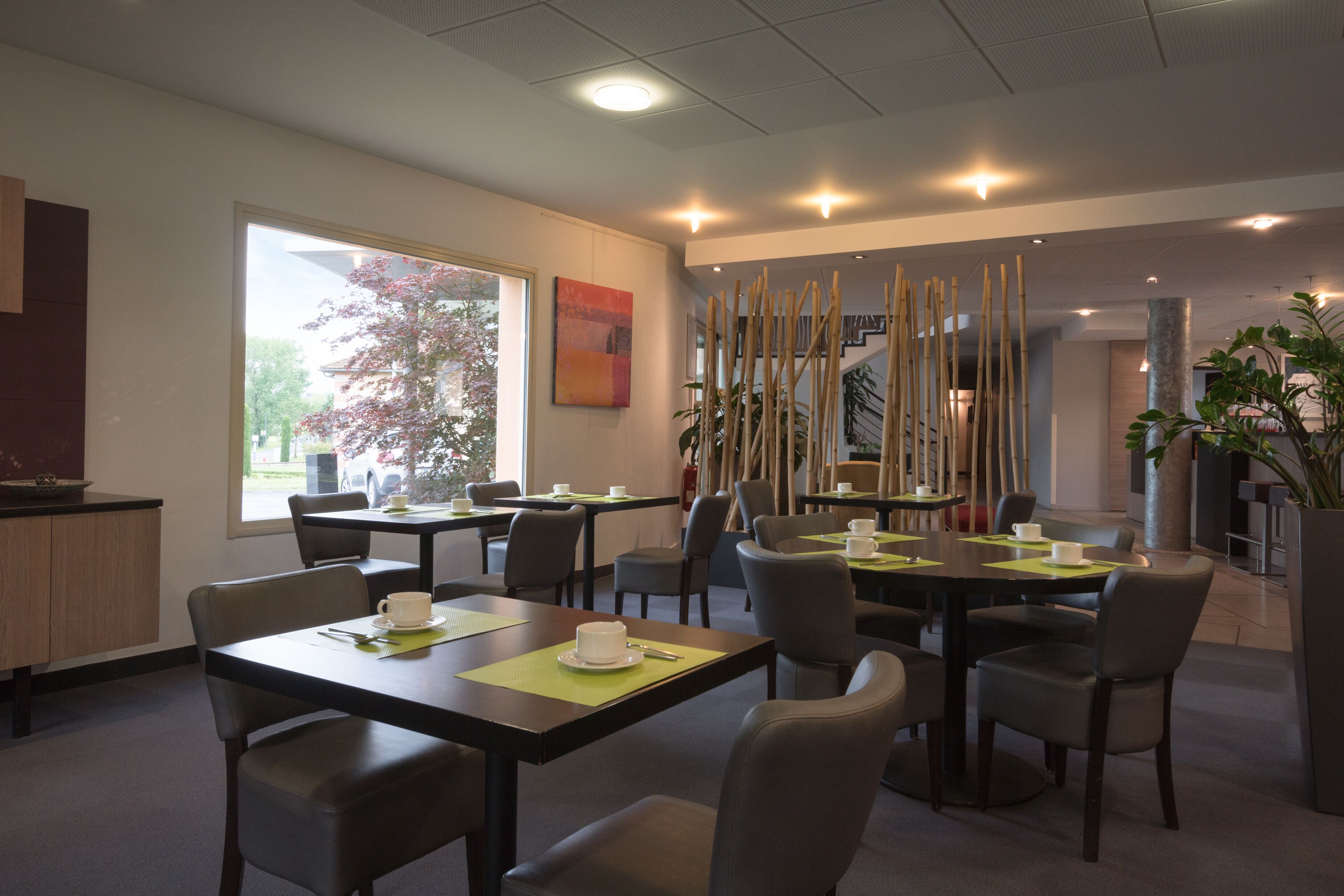 Un comedor moderno con mesas y sillas, un ventanal con vistas a árboles y un panel decorativo de bambú.