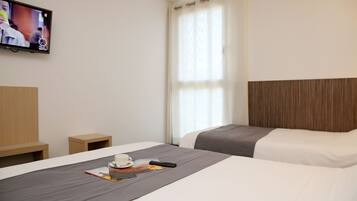 A hotel room with two beds, a TV mounted on the wall, a small table with a cup and a book, and a window with curtains.