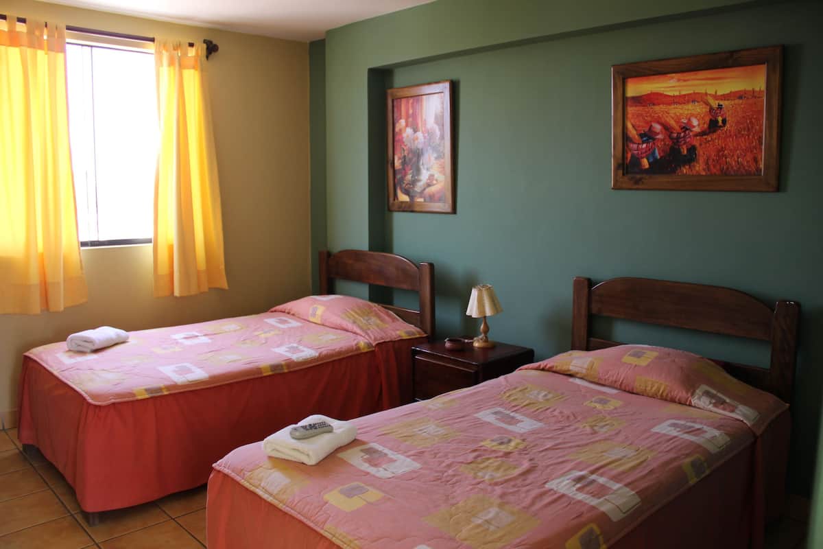 A room with two single beds, a nightstand with a lamp, a painting on the wall, and a window with curtains.