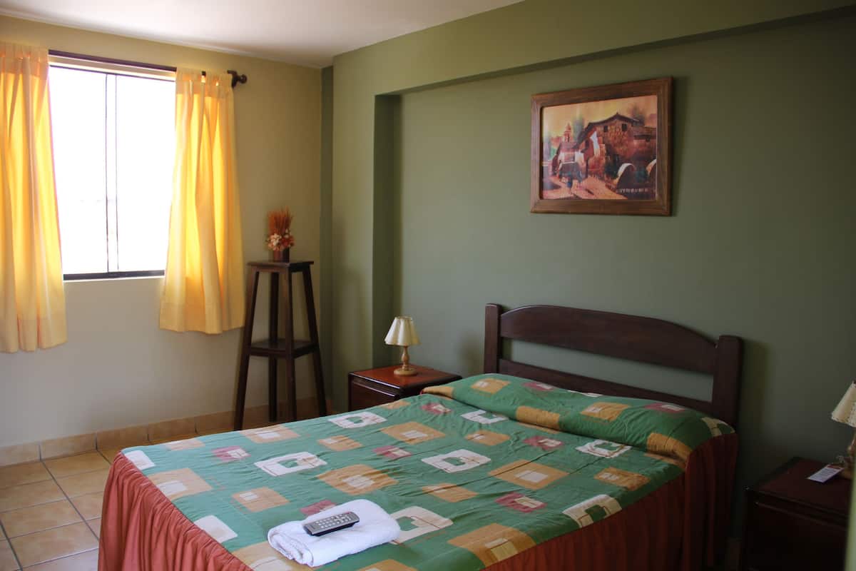 A bedroom with a bed, a nightstand, a lamp, a window with curtains, and a painting on the wall.