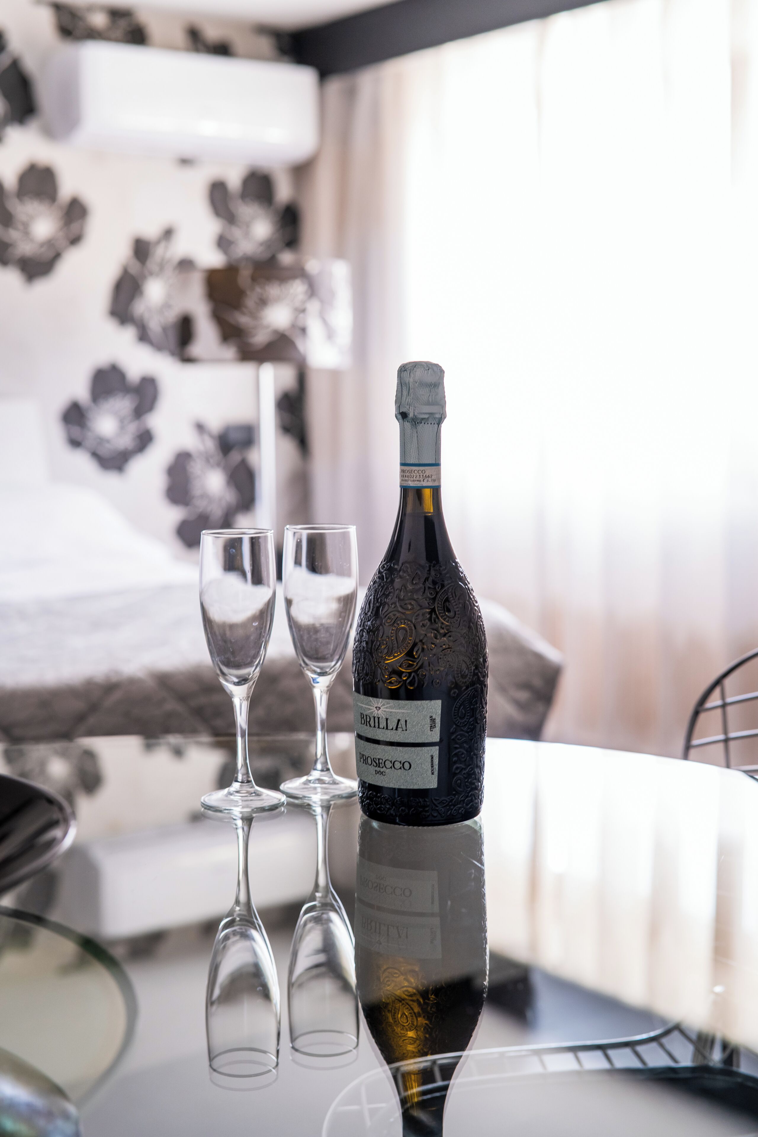 Una bottiglia di vino BRILLAI e due bicchieri su una superficie riflettente in una stanza con carta da parati a motivi floreali.