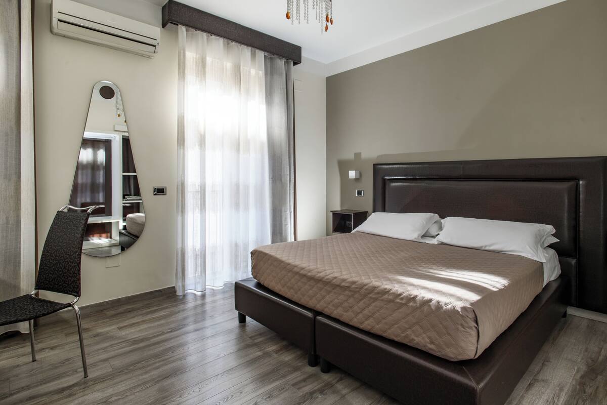 Une chambre d’hôtel avec un lit, une chaise, un bureau, un miroir et une fenêtre avec des rideaux.