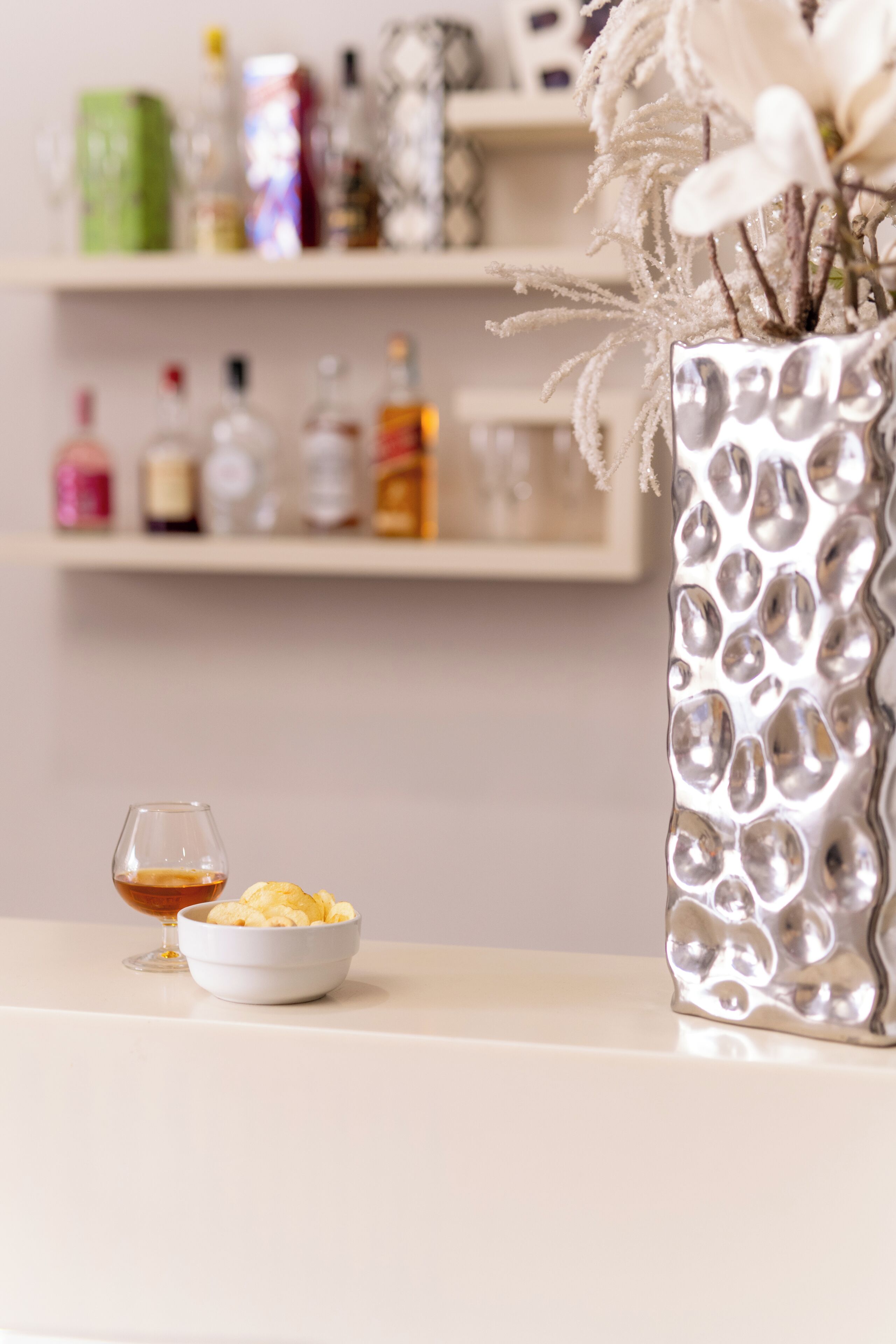 A table with a bowl of chips and a glass of drink, a decorative vase with flowers, and shelves with various bottles in the background.