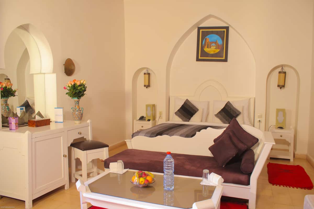 A bedroom with a bed, a desk, a chair, a table with a fruit bowl and a water bottle, and a red rug.