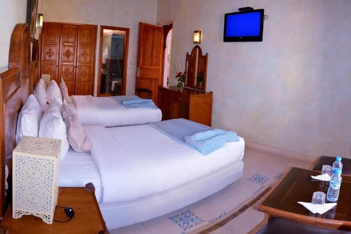 A hotel room with two beds, a television, a desk with a chair, and a small table with a bottle and glasses.