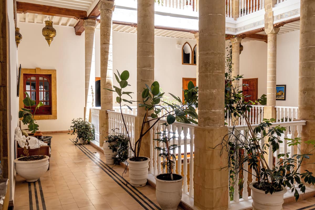 A spacious corridor with large columns, potted plants, and a wooden railing.