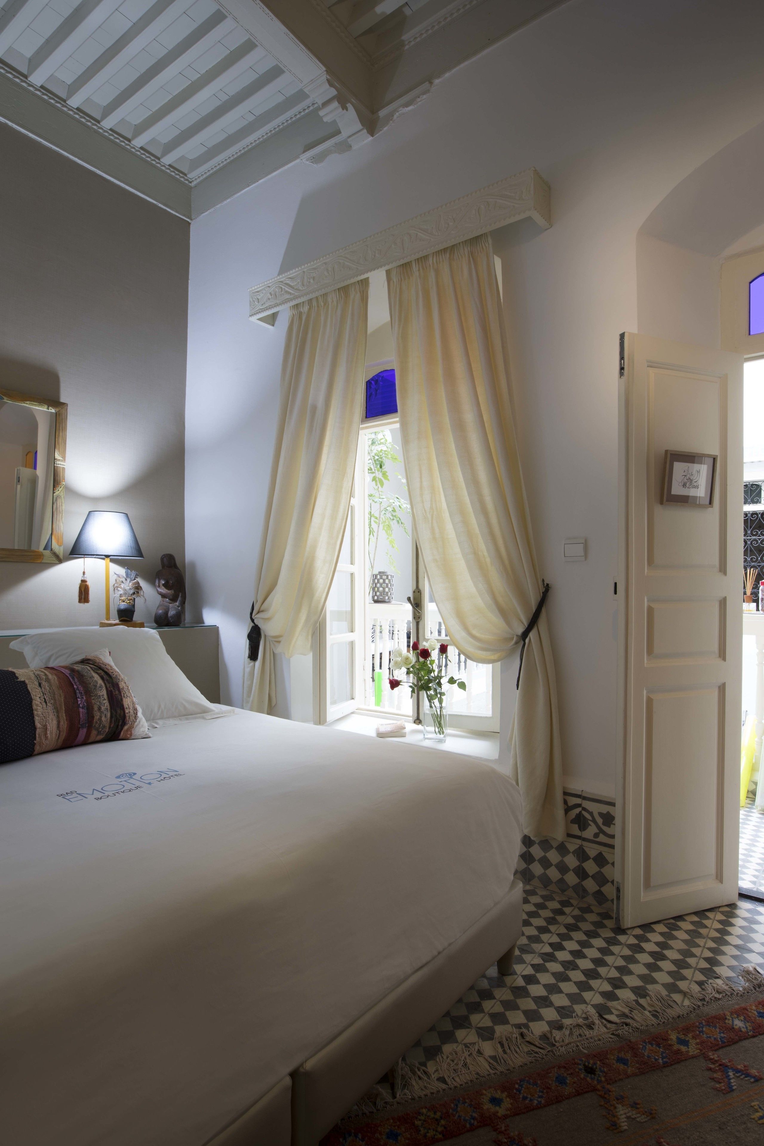 A bedroom with a bed, a nightstand, a lamp, a window with curtains, and a door leading to another room.
