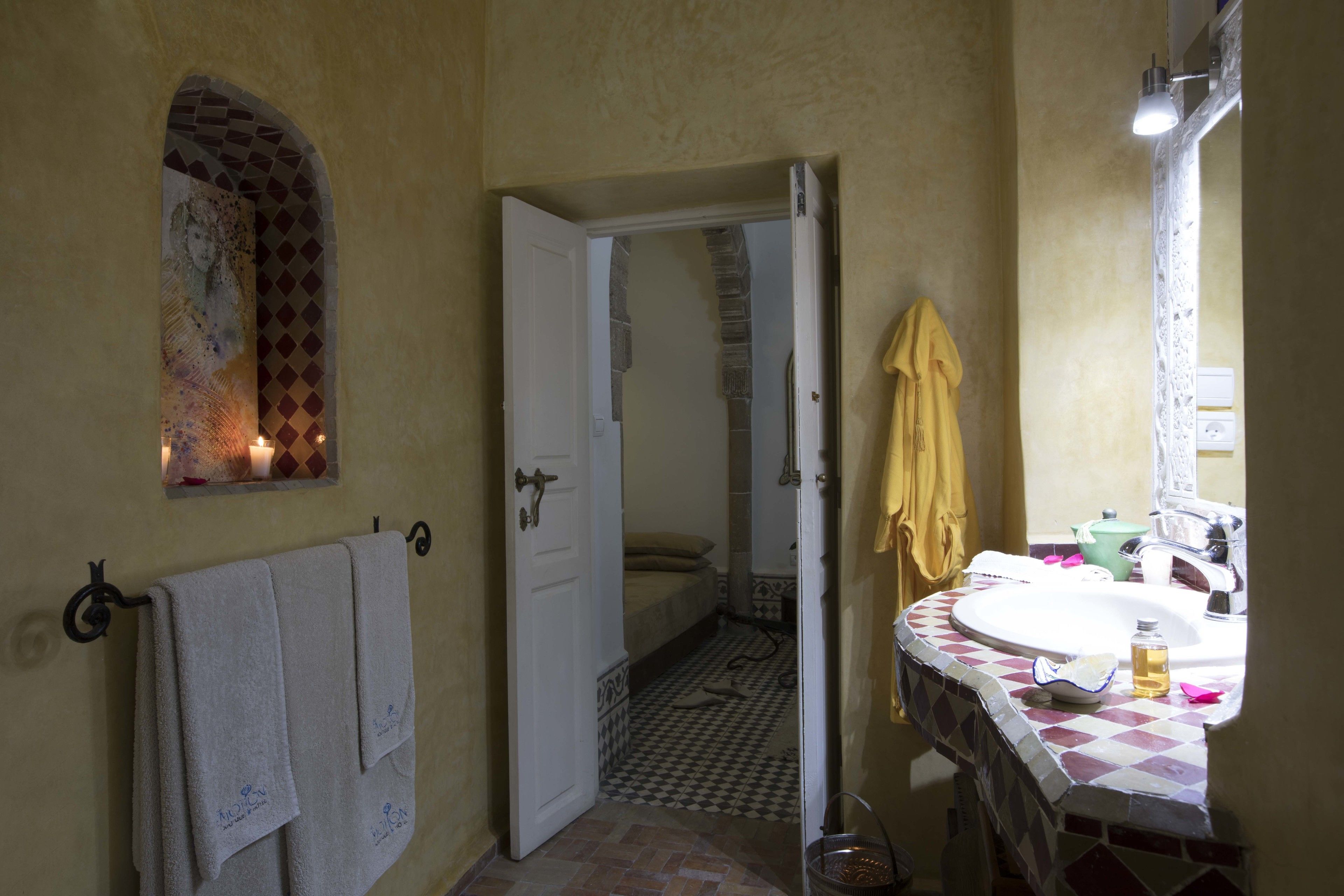 A bathroom with a tiled floor, a sink with a mirror, a towel rack, and a door leading to another room.