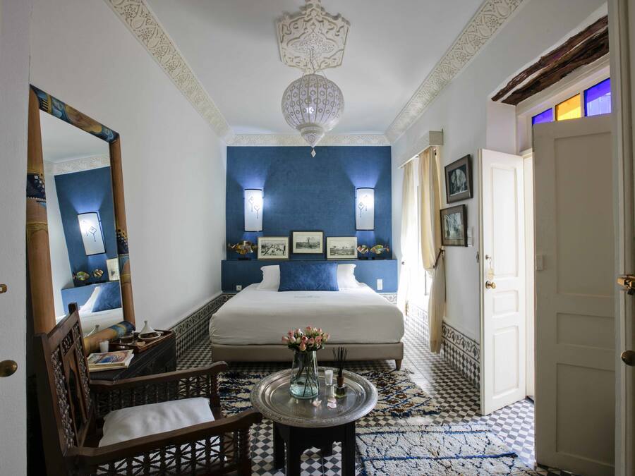 A hotel room with a large bed, a sitting area with a chair, a small table with a vase, and a decorative ceiling light.