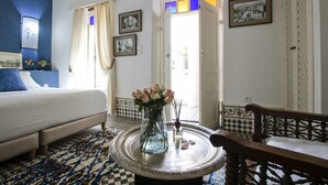 Une chambre à coucher avec un lit, une table ronde ornée d’un vase de fleurs, un banc en bois et une porte donnant sur un balcon.