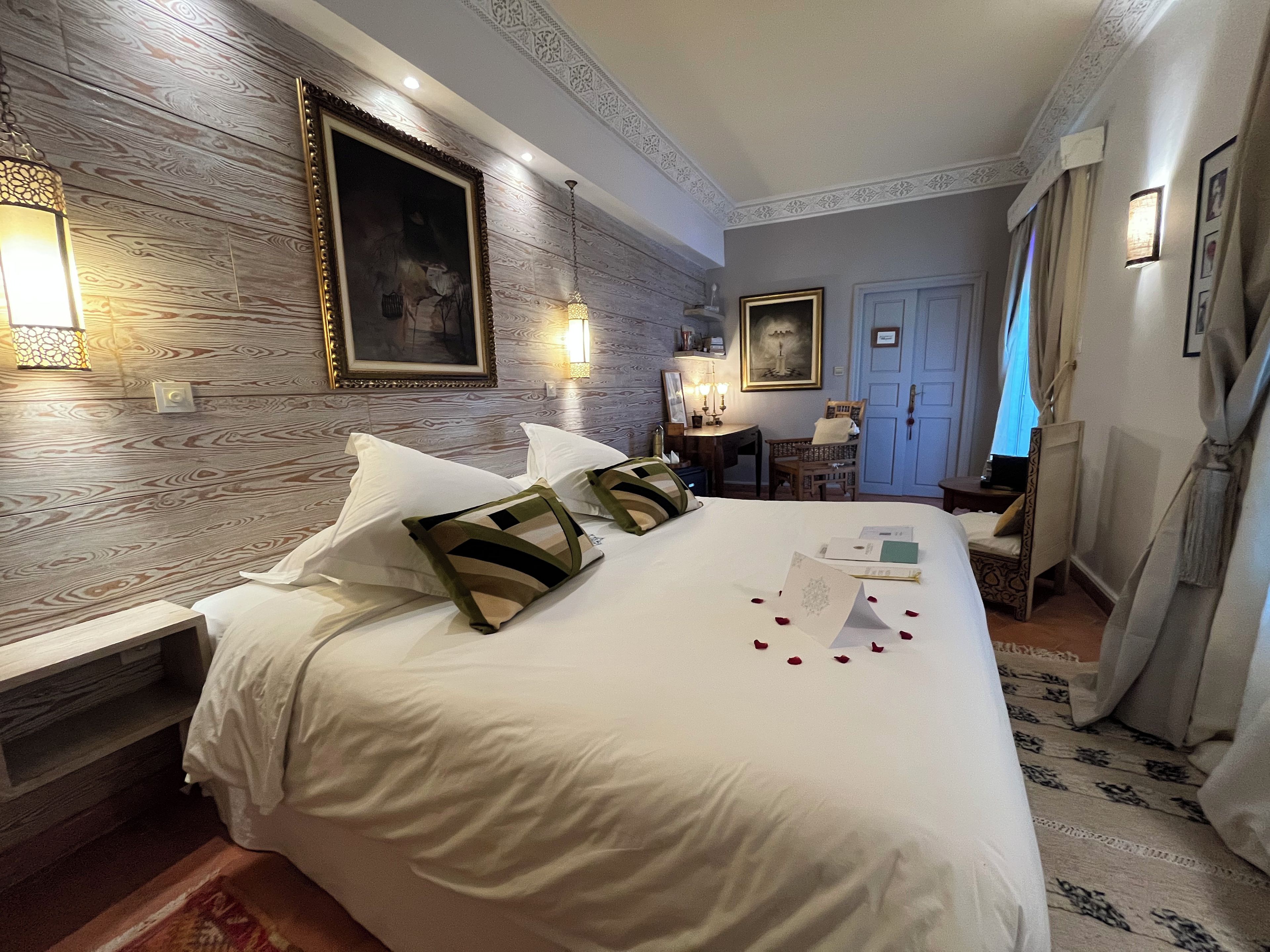 Une chambre d’hôtel avec un grand lit, une tête de lit en bois, un tableau, un bureau avec une lampe et une porte avec des rideaux.