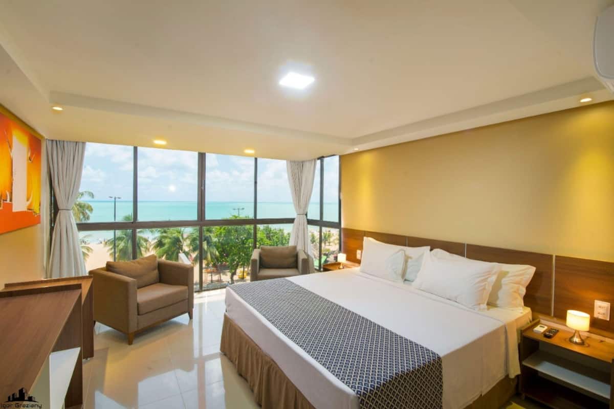 A hotel room with a large bed, two armchairs, a view of the ocean, and a cityscape.