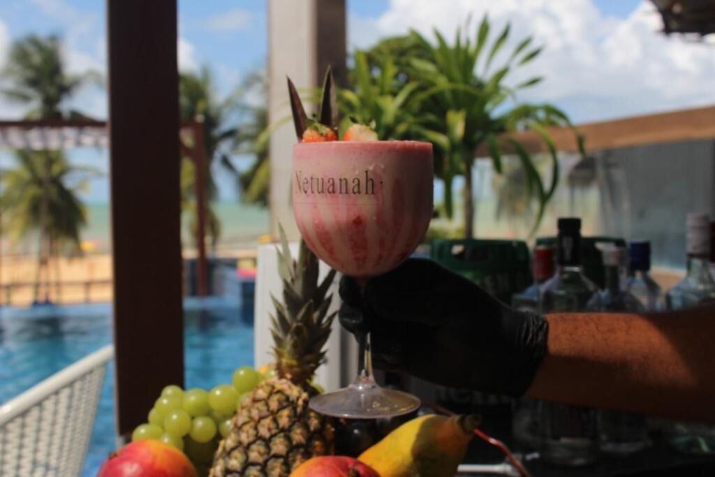 Un cóctel en un vaso con la etiqueta 'Netuanah', adornado con fruta tropical, piña y otras frutas, sobre una barra con vistas a una piscina.
