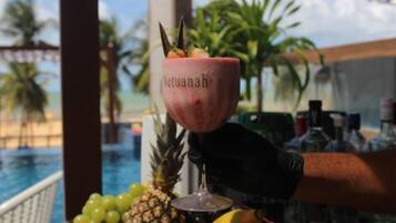 Un cóctel en un vaso con la etiqueta 'Netuanah', adornado con fruta tropical, piña y otras frutas, sobre una barra con vistas a una piscina.