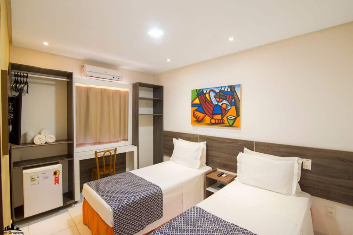A hotel room with two beds, a desk, a chair, an air conditioner, a mini-fridge, and a colorful painting on the wall.