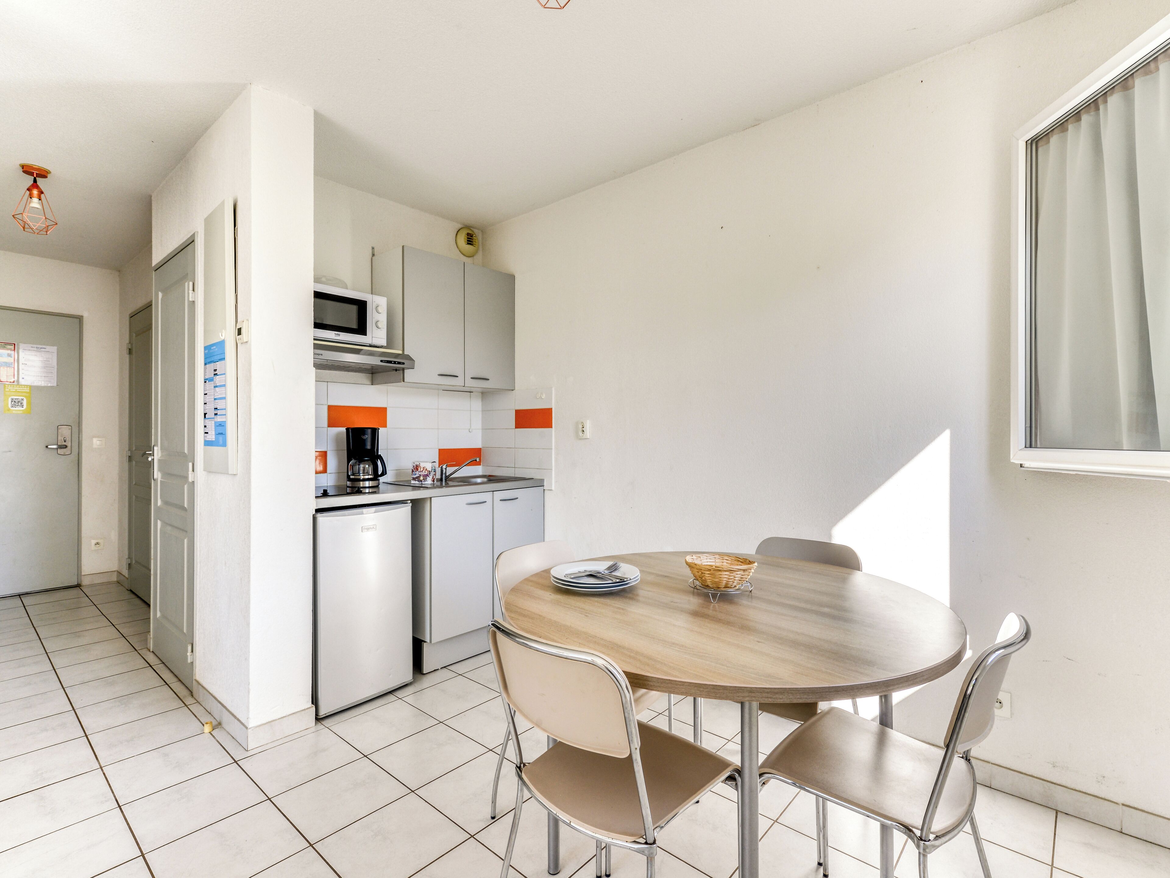 Cocina compacta con microondas, cafetera y lavaplatos. Se observa una mesa de comedor con sillas y un ventanal.