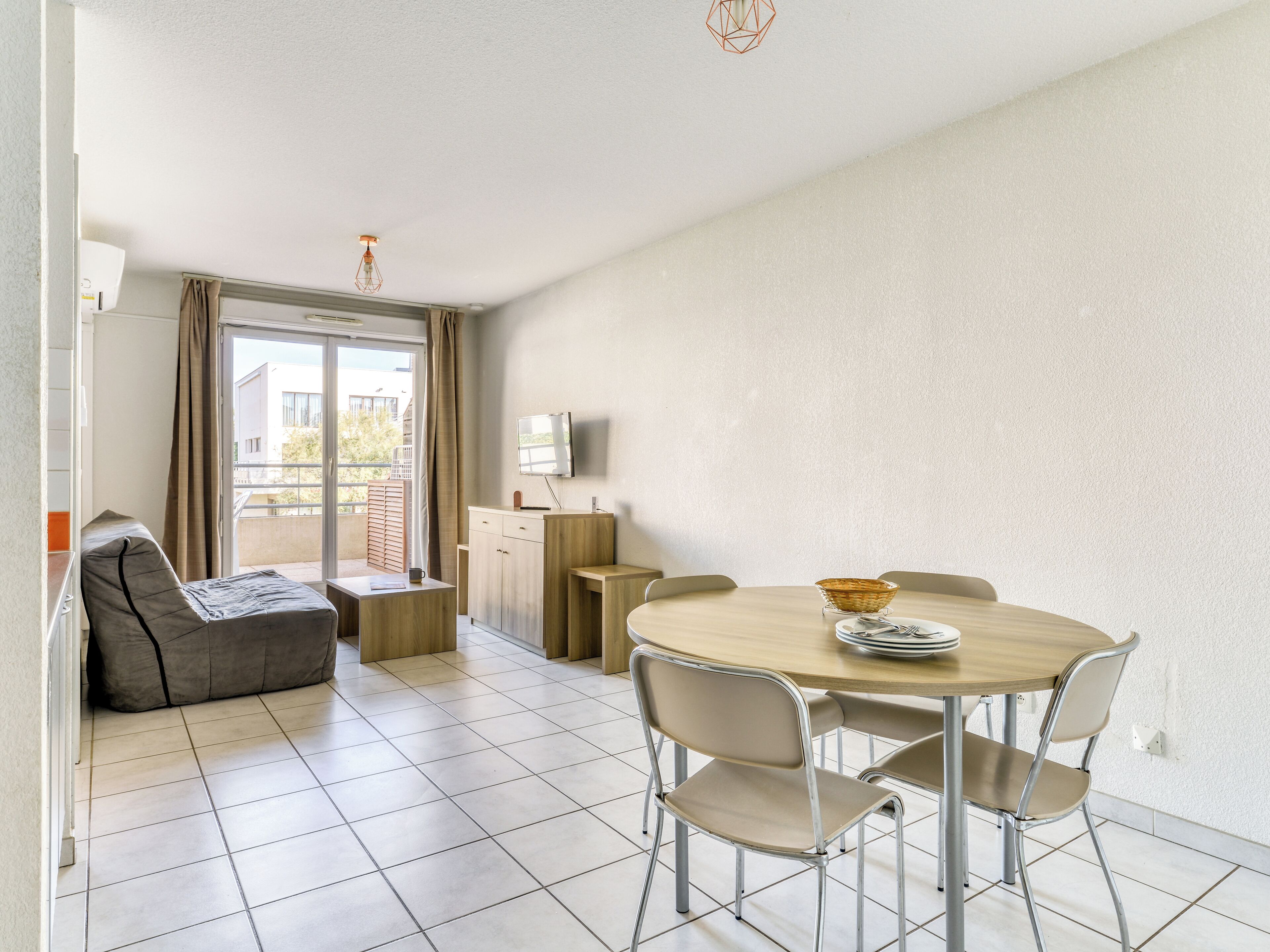 Una sala de estar moderna con un sofá, una mesa de comedor con sillas, un televisor montado en la pared y un balcón con vistas a edificios.