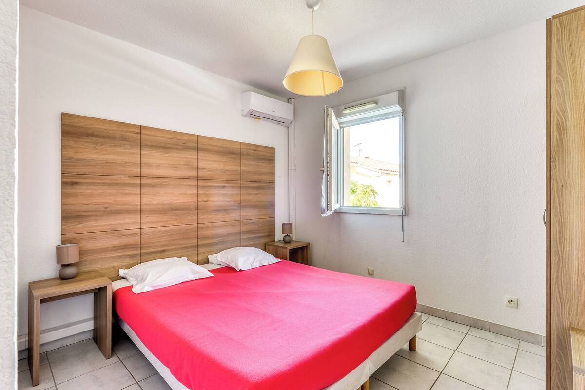 A bedroom with a wooden headboard, a red bedspread, two bedside tables with lamps, a window with a view, and an air conditioning unit.