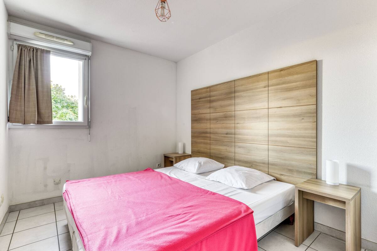 A bedroom with a wooden headboard, a bed with white linens, a pink blanket, and a window with curtains.