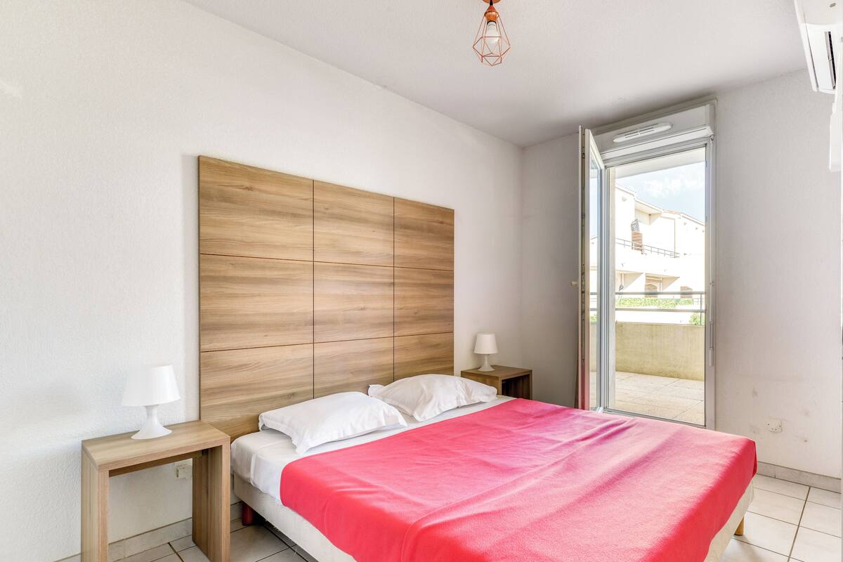 A bedroom with a wooden headboard, a bed with white and red bedding, two bedside tables with lamps, and a sliding glass door leading to an outdoor area.