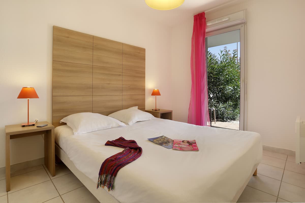A neatly made bed with white linens, two bedside lamps, and a window with a view of greenery.
