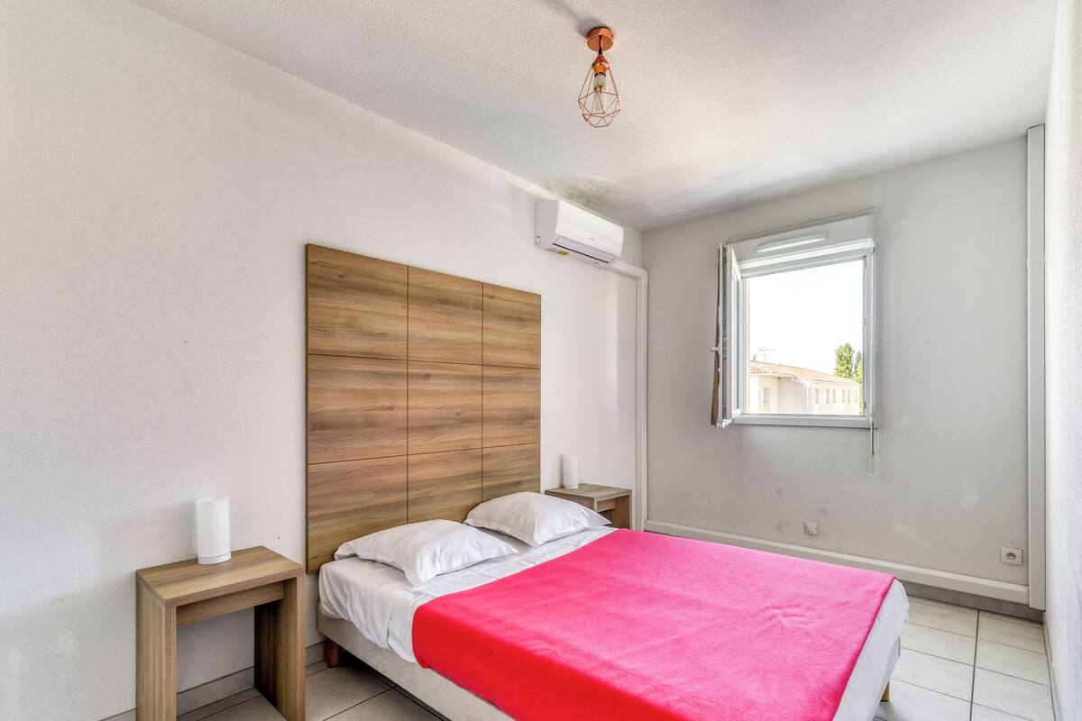 A bedroom with a wooden headboard, a single bed with white bedding and a red blanket, a bedside table with a white lamp, and a window with a view of buildings outside.