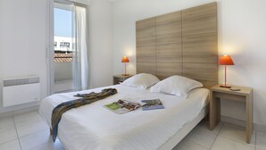 A modern bedroom with a large bed, two bedside tables with lamps, a window with curtains, and a view of buildings outside. - Vacancéole - Cap Camargue (Le Grau-Du-Roi)